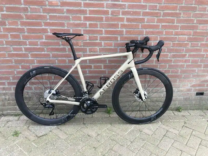 Aeross Endurance endurance racefiets in mocha met carbon wielen, zijaanzicht tegen een bakstenen muur